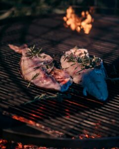 Close-up of fish slices being grilled with herbs over an open flame, highlighting outdoor cooking.